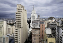 A imagem mostra uma vista panorâmica da cidade de São Paulo, com vários edifícios altos e modernos. O céu está nublado, criando uma atmosfera dramática. No centro, destaca-se um edifício icônico com uma bandeira no topo. A cidade é densamente povoada, com uma grande quantidade de prédios e estruturas urbanas visíveis ao fundo.