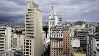 A imagem mostra uma vista panorâmica da cidade de São Paulo, com vários edifícios altos e modernos. O céu está nublado, criando uma atmosfera dramática. No centro, destaca-se um edifício icônico com uma bandeira no topo. A cidade é densamente povoada, com uma grande quantidade de prédios e estruturas urbanas visíveis ao fundo.