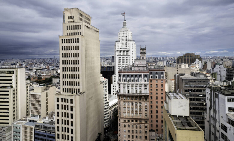 A imagem mostra uma vista panorâmica da cidade de São Paulo, com vários edifícios altos e modernos. O céu está nublado, criando uma atmosfera dramática. No centro, destaca-se um edifício icônico com uma bandeira no topo. A cidade é densamente povoada, com uma grande quantidade de prédios e estruturas urbanas visíveis ao fundo.