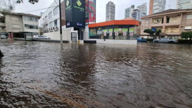 A imagem mostra uma rua inundada em uma área urbana, com água cobrindo parcialmente os veículos estacionados e a calçada. No fundo, há prédios altos e um comércio com uma fachada colorida. O céu está nublado, indicando um clima chuvoso.
