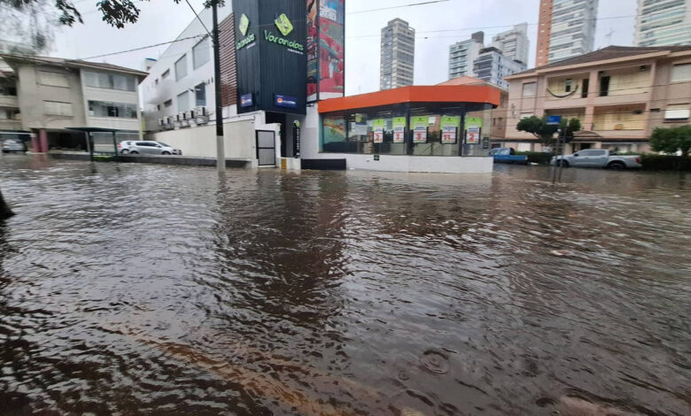 A imagem mostra uma rua inundada em uma área urbana, com água cobrindo parcialmente os veículos estacionados e a calçada. No fundo, há prédios altos e um comércio com uma fachada colorida. O céu está nublado, indicando um clima chuvoso.