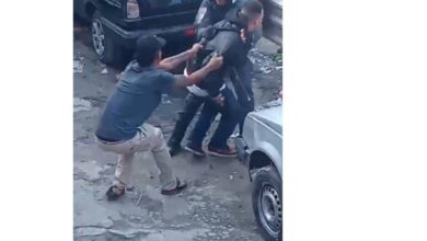A imagem mostra um grupo de pessoas em uma situação de conflito em uma área urbana. Algumas pessoas estão se segurando e parecem estar em uma discussão ou briga. Ao fundo, há vários carros estacionados. O ambiente parece ser uma rua ou um estacionamento.