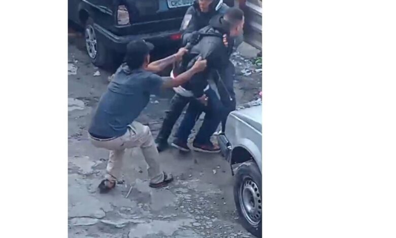 A imagem mostra um grupo de pessoas em uma situação de conflito em uma área urbana. Algumas pessoas estão se segurando e parecem estar em uma discussão ou briga. Ao fundo, há vários carros estacionados. O ambiente parece ser uma rua ou um estacionamento.