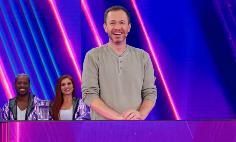 Um apresentador sorridente está em pé atrás de uma mesa em um cenário colorido e moderno. Ele usa uma camiseta de manga longa cinza e está cercado por um grupo de pessoas que estão sentadas atrás dele, todas vestindo jaquetas brilhantes. O fundo é iluminado com luzes em tons de roxo e azul, e há um texto visível que diz