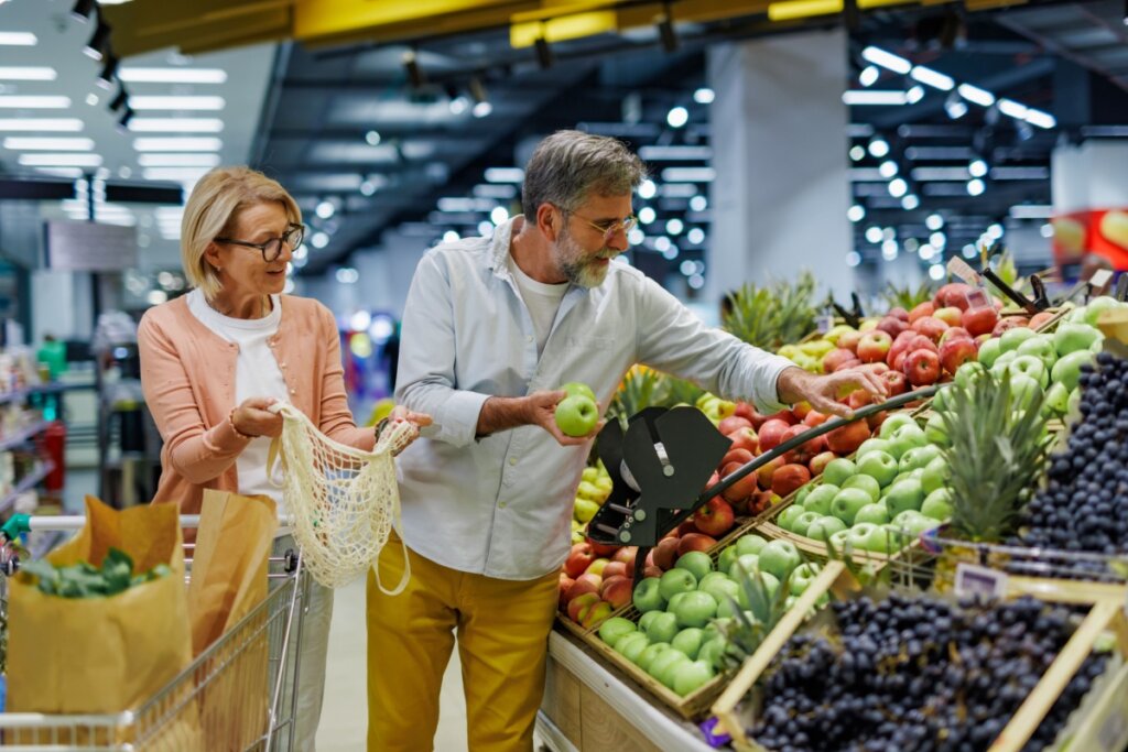 Um casal de meia-idade faz compras na seção de hortifrúti de um supermercado iluminado. O homem, com barba grisalha e camisa clara, segura uma maçã verde enquanto escolhe outras frutas em uma bancada repleta de maçãs vermelhas, verdes e uvas. À frente deles, um carrinho de compras com sacos de papel cheios de mantimentos.