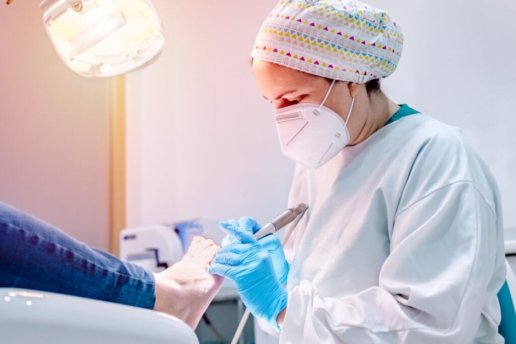 Médica usando touca, máscara e uniforme branco realizando procedimento em unha de paciente