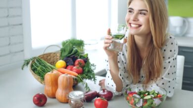 Mulher loira sorrindo e com camisa branca de bolinha, segurando copo de vidro com água e apoiada em bancada com alimentos vegetais e pote com salada