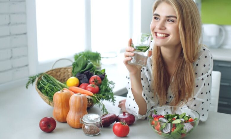 Mulher loira sorrindo e com camisa branca de bolinha, segurando copo de vidro com água e apoiada em bancada com alimentos vegetais e pote com salada
