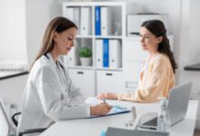 Médica com cabelo liso, solto usando jaleco branco fazendo anotações em prancheta e paciente sentada de frente pra médica com o cabelo liso semi preso e usando camisa de manga longa laranja-clara