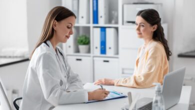 Médica com cabelo liso, solto usando jaleco branco fazendo anotações em prancheta e paciente sentada de frente pra médica com o cabelo liso semi preso e usando camisa de manga longa laranja-clara