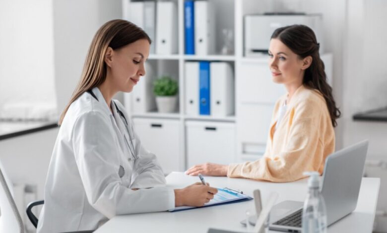 Médica com cabelo liso, solto usando jaleco branco fazendo anotações em prancheta e paciente sentada de frente pra médica com o cabelo liso semi preso e usando camisa de manga longa laranja-clara
