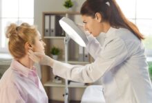 Dermatologista analisando pele do rosto de paciente em consultório, a médica está com o cabelo preso em rabo de cavalo, usando jaleco e luvas brancas e a paciente tem o cabelo loiro preso em coque e está usando camisa social rosa