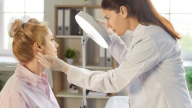 Dermatologista analisando pele do rosto de paciente em consultório, a médica está com o cabelo preso em rabo de cavalo, usando jaleco e luvas brancas e a paciente tem o cabelo loiro preso em coque e está usando camisa social rosa