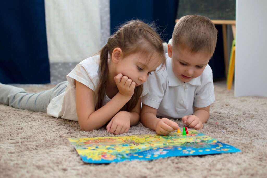 Os jogos e atividades lúdicas estimulam imaginação, raciocínio e adaptação Duas crianças deitadas em tapete brincando com jogo de tabuleiro