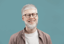 Homem albino com cabelo curto e barba, usando camiseta branca, camisa em tom de marrom claro e óculos preto