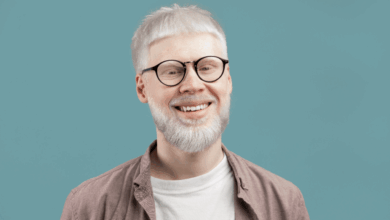 Homem albino com cabelo curto e barba, usando camiseta branca, camisa em tom de marrom claro e óculos preto