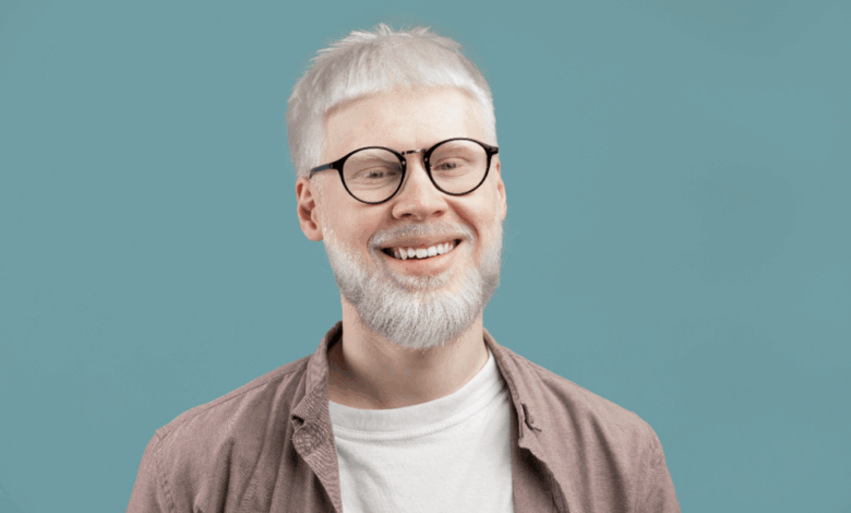Homem albino com cabelo curto e barba, usando camiseta branca, camisa em tom de marrom claro e óculos preto